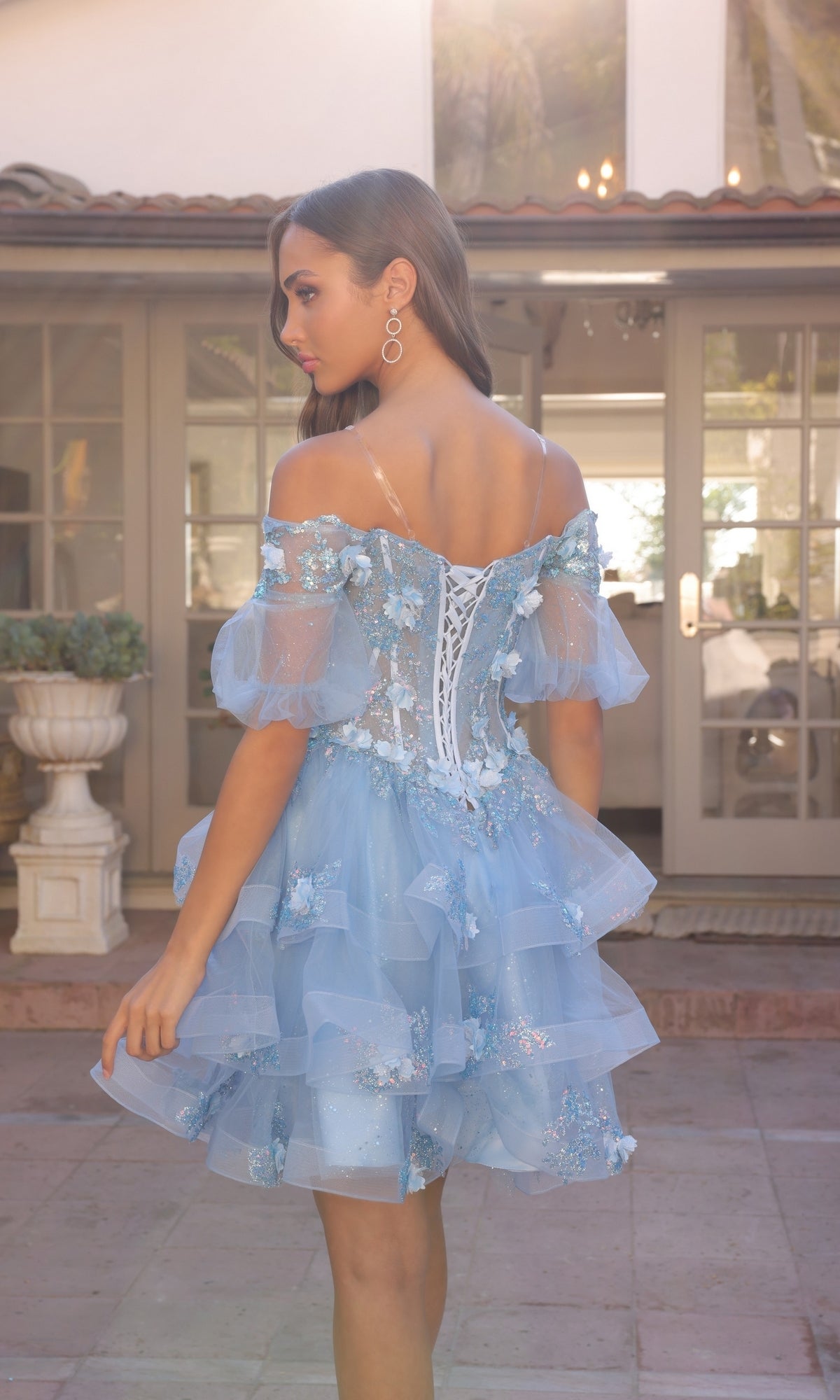 A woman wears the Nox Anabel Removable-Skirt Formal Ball Gown H1360, featuring floral embroidery and ruffled layers, standing outdoors with her back to the camera.